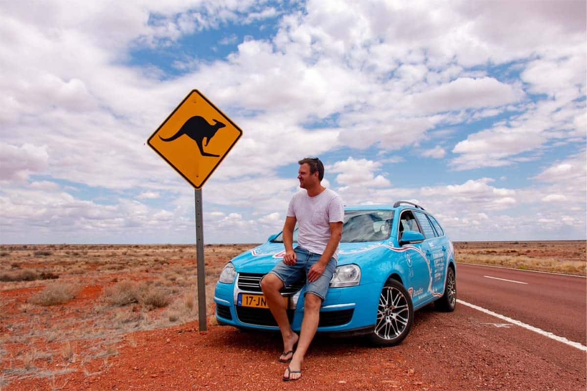 Wiebe Wakker sitting on the bonet of his electic car on a road in Australia next to a kangaroo warning sign.