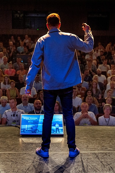 Wiebe Wakker from the back, he is facing an audience while he is speaking on stage in a theatre.