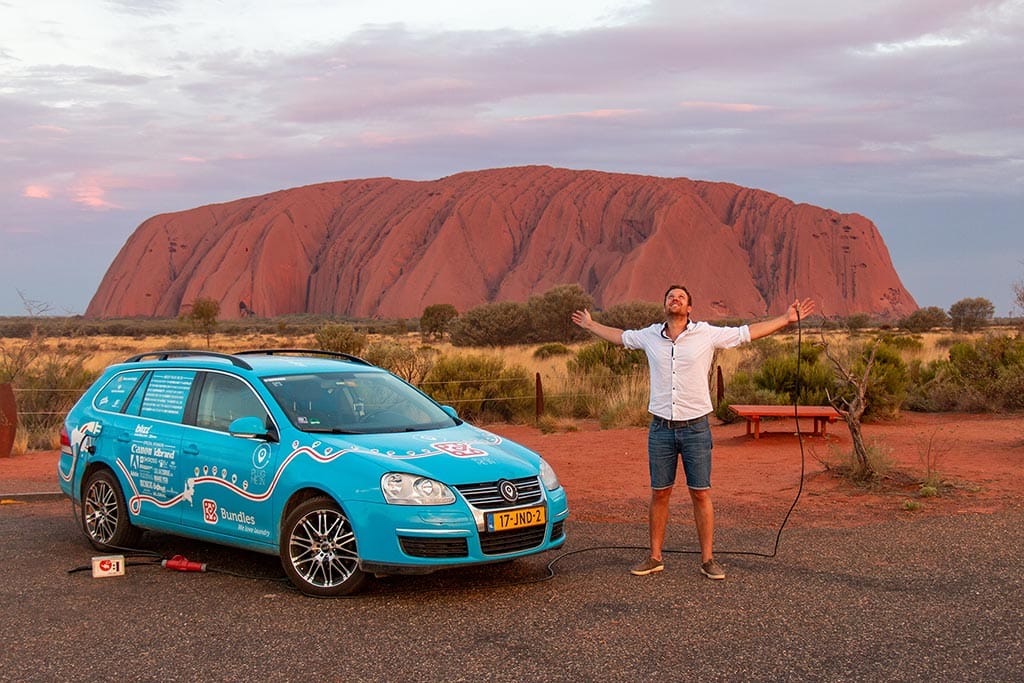 Wiebe Wakker standing next to his Blue Bandit electric car, in front of Uluru, Australia.
